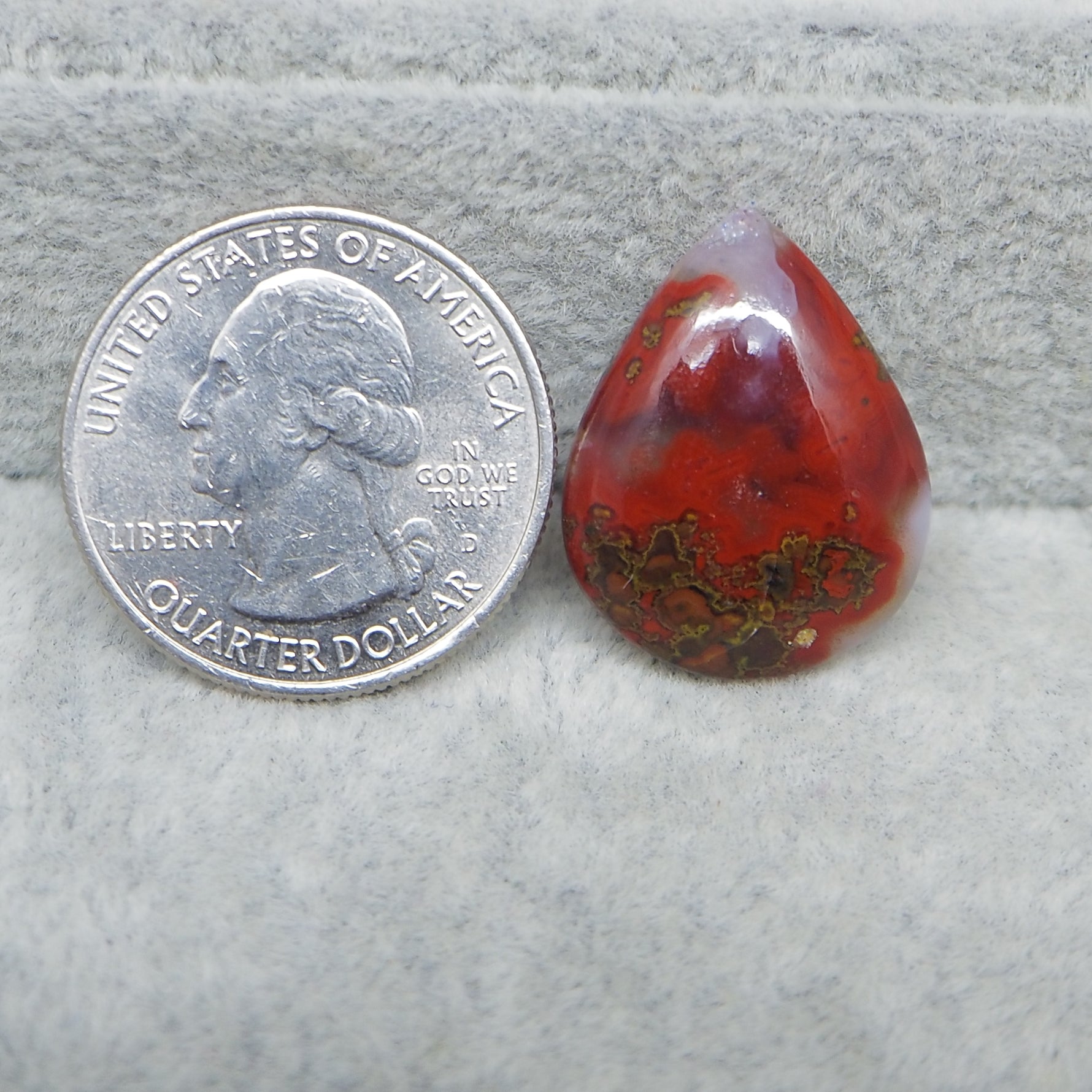 Red teardrop-shaped stone next to a U.S. quarter dollar on a gray surface