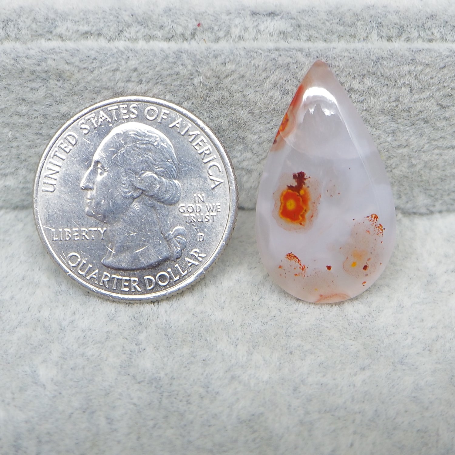 Teardrop-shaped agate stone next to a U.S. quarter dollar coin on a textured gray surface.