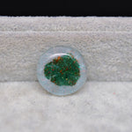Round glass cabochon with green and red speckles on a textured surface