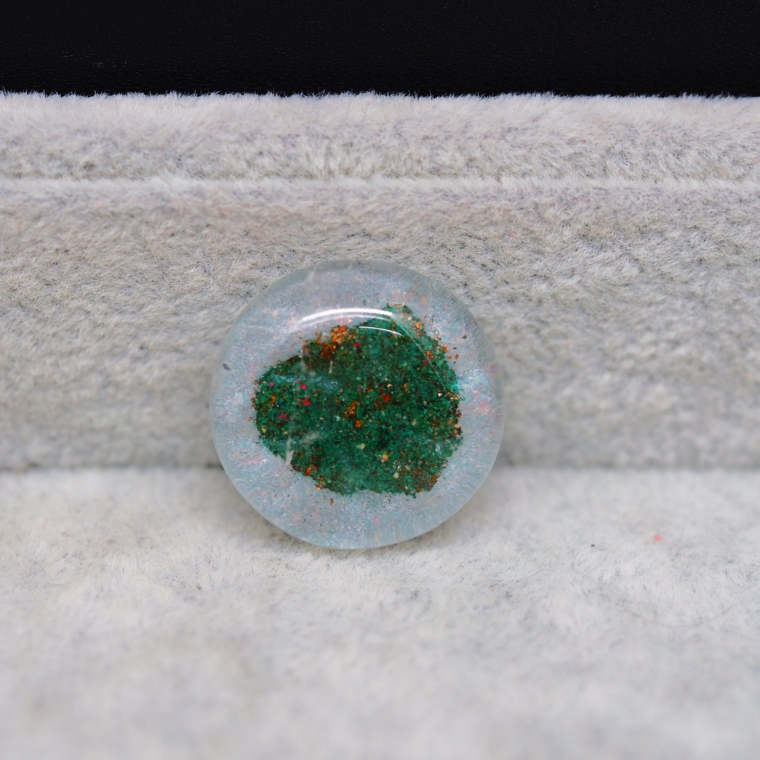 Round glass cabochon with green and red speckles on a textured surface