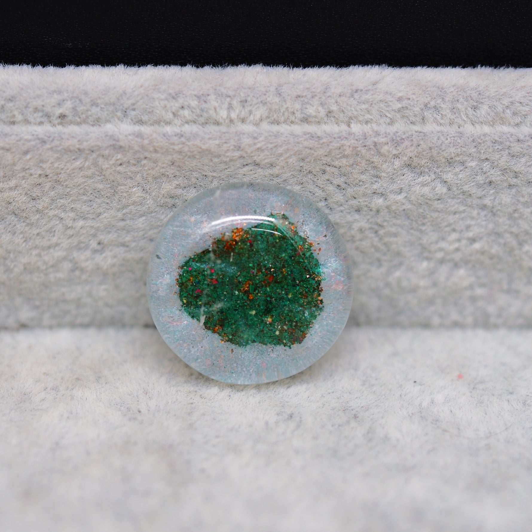 Round glass cabochon with green and red speckles on a textured surface